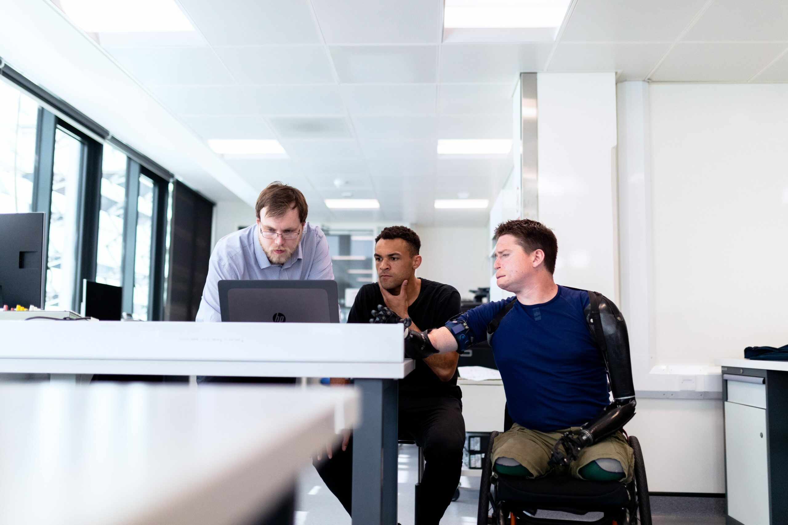 Three men sit around a desk looking at a computer screen. One of the men is in a wheelchair and is paraplegic.
